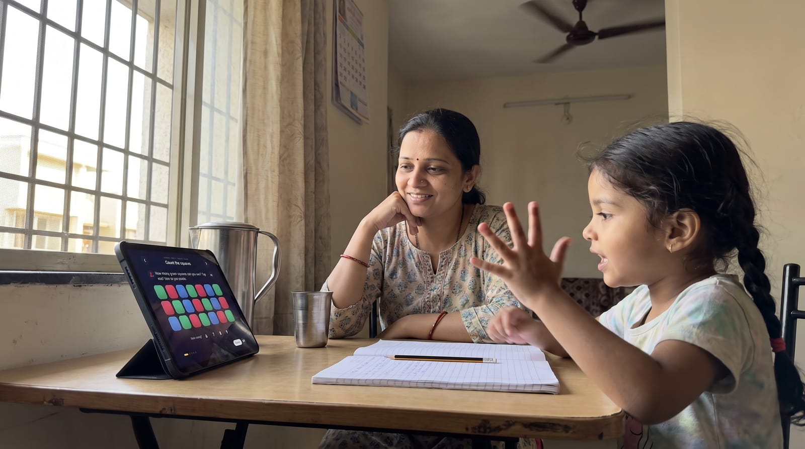 A mother and her five-year-old daughter at a dining table, the girl answering a counting question on a tablet